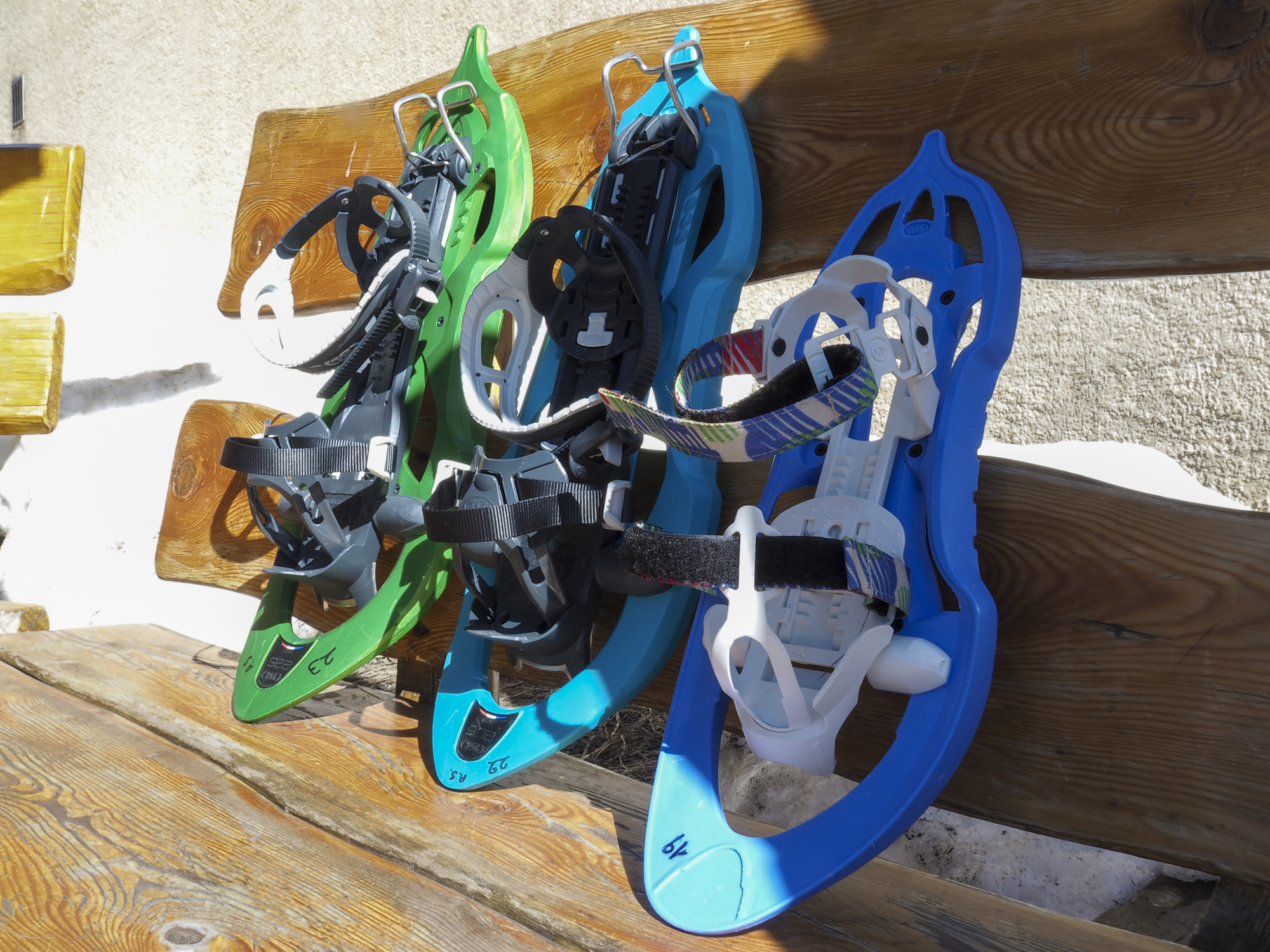Three pairs of colorful snowshoes, green, turquoise, and blue, leaning against a wooden bench on a sunny day in the mountains.